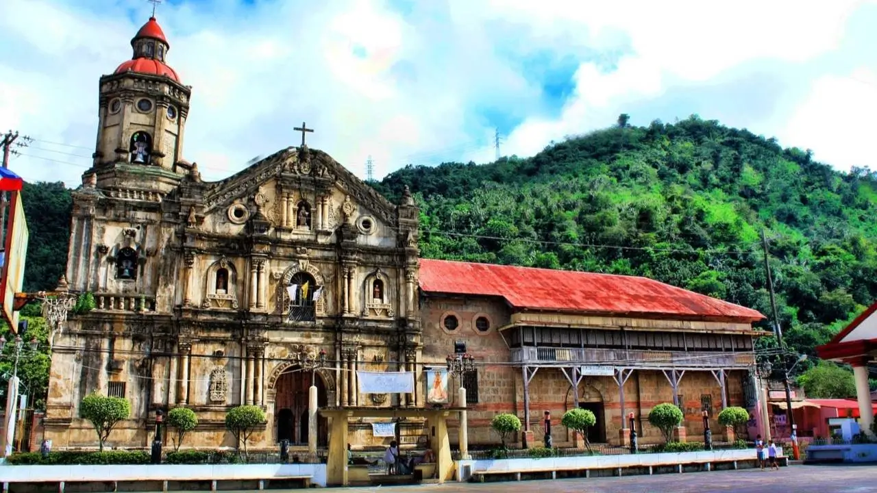 Antigua iglesia con fachada ornamentada, torre con cúpula roja y cruz, rodeada de árboles y colinas verdes bajo un cielo parcialmente nublado