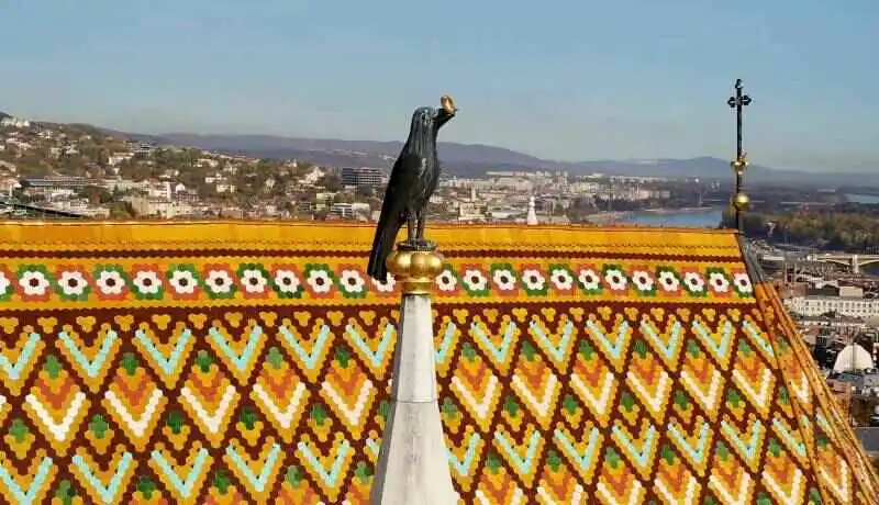 # Descripción para texto alternativo (alt text)  **Escultura de un cuervo negro con un anillo dorado en el pico, situada sobre una aguja blanca con remate dorado, frente a un tejado decorado con tejas de colores en patrones geométricos, con una ciudad y colinas al fondo.