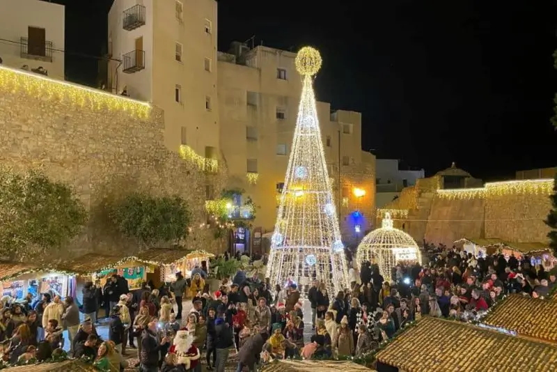 La imagen muestra un mercado navideño en Peñíscola, España, que es un evento popular con un gran árbol de Navidad iluminado y puestos llenos de gente.