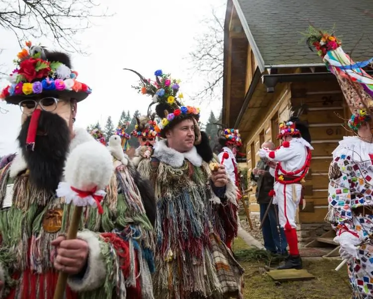 La imagen retrata una celebración folclórica en un entorno rural, probablemente vinculada al carnaval tradicional de la región de Vysočina, en la República Checa. Los participantes visten trajes elaborados hechos de lana multicolor, pieles y telas, con tocados ornamentales decorados con flores artificiales, plumas y otros elementos festivos. Algunos llevan máscaras o cubren sus rostros, y uno sostiene un bastón decorado, lo que sugiere un papel ritual o simbólico dentro del desfile