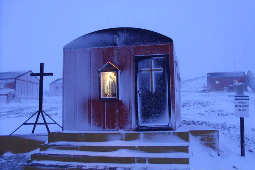 Esta fotografía captura la capilla san francisco de asis en la Antártida, un pequeño edificio rojo con una cruz en la cima, rodeado de nieve y hielo. Situada en la costa argentina, esta estación de investigación sirve como un centro científico clave para el estudio del entorno y el clima del continente.