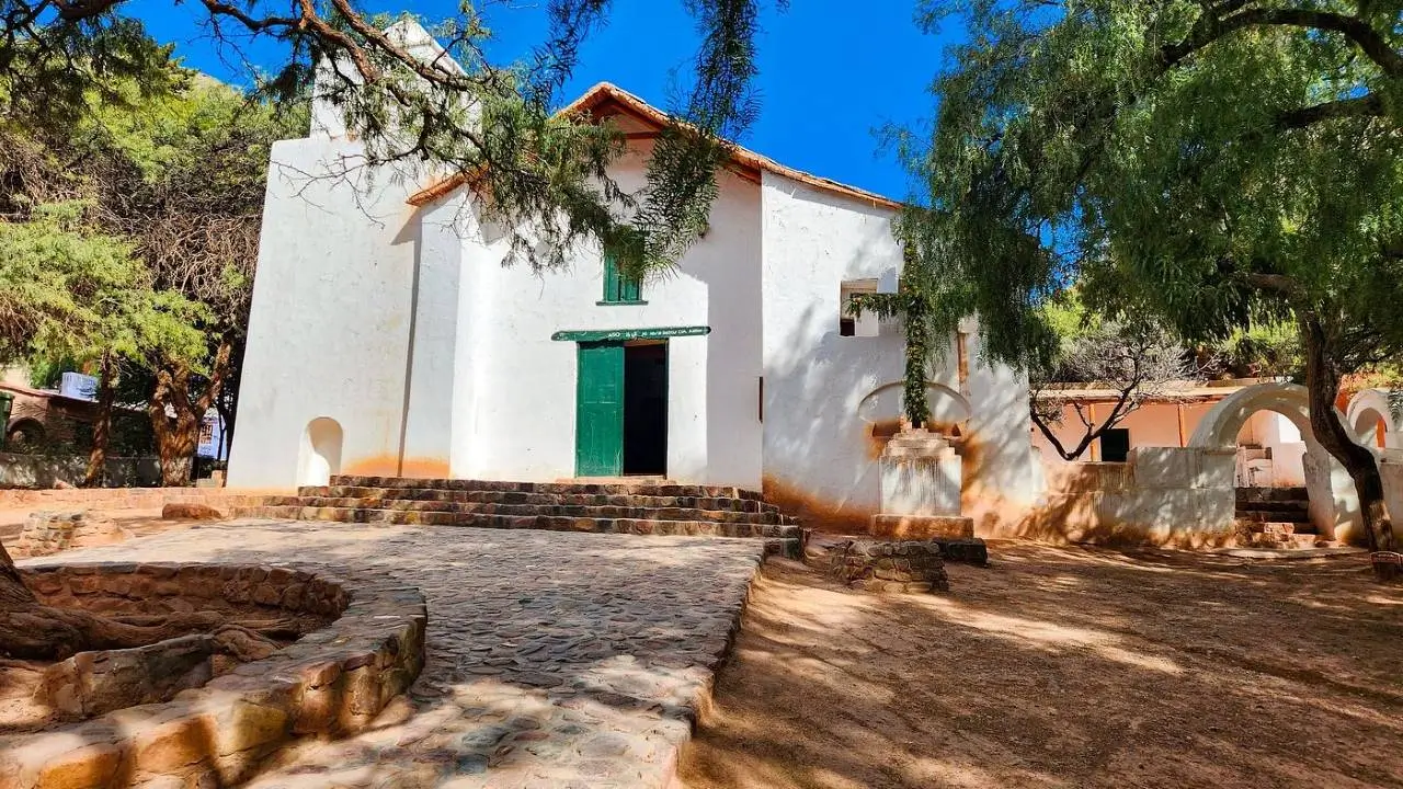 Esta imagen muestra la Iglesia Santa Rosa de Lima en Purmamarca, Jujuy. Es un Monumento Histórico Nacional construido con muros de adobe y techos de cardón. La fachada blanca contrasta notablemente con el paisaje colorido de los cerros de la zona. turismo religioso en jujuy