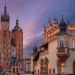 Esta fotografía captura la Basílica de Santa María y la Lonja de los Paños de Cracovia bañadas por el cálido resplandor de un sol poniente sobre la histórica plaza del Rynek Główny. La escena muestra la mezcla de arquitectura gótica y renacentista que caracteriza el Casco Antiguo de Cracovia, turismo religioso en Polonia.