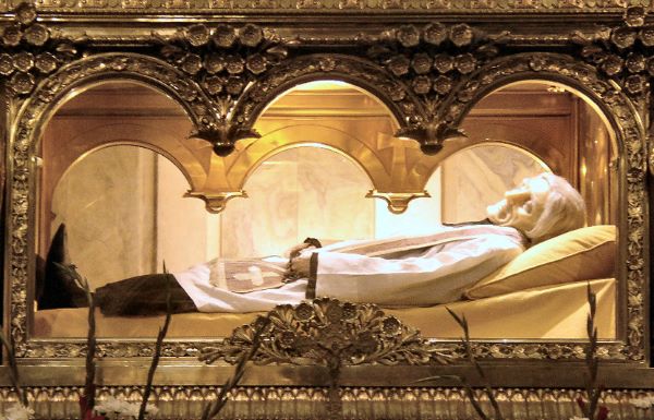 La imagen muestra el cuerpo incorrupto de San Juan María Vianney (el Cura de Ars), expuesto en una urna de cristal y bronce dentro de la basílica de Ars-sur-Formans, en Francia