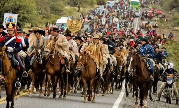 La imagen muestra una tradicional cabalgata de la fe gauchesca sobre una ruta en Argentina, donde numerosos jinetes vestidos con ponchos y atuendos típicos avanzan en procesión o desfile acompañados de banderas y estandartes