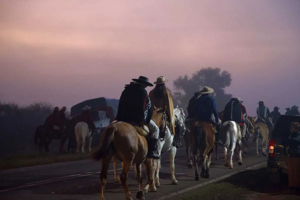La imagen muestra una columna de jinetes o gauchos con vestimenta tradicional y ponchos a caballo transitando por una ruta pavimentada durante las primeras horas de la mañana o el atardecer, bajo un cielo brumoso y rosáceo