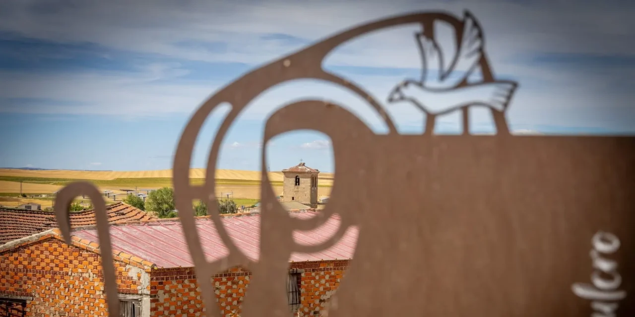 La imagen muestra una escultura o silueta metálica decorativa en primer plano, con un diseño que parece representar la figura estilizada de un peregrino y un ave, enmarcando un paisaje rural castellano con casas de ladrillo y una torre al fondo,