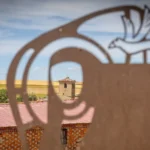 La imagen muestra una escultura o silueta metálica decorativa en primer plano, con un diseño que parece representar la figura estilizada de un peregrino y un ave, enmarcando un paisaje rural castellano con casas de ladrillo y una torre al fondo,