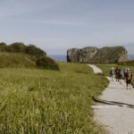 La imagen muestra a un grupo de peregrinos caminando por un sendero verde junto a la costa, marcado por un poste de madera con una señal azul y la tradicional concha de vieira del Camino de Santiago