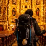 La imagen muestra a un peregrino con una mochila y una vieira tradicional de utilería o símbolo del Camino de Santiago dentro de una iglesia ornamentada. Credito Peter Amende / Unsplash