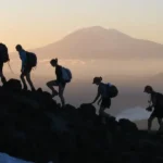 La imagen muestra una silueta de un grupo de excursionistas (cinco personas con mochilas y bastones de trekking) subiendo por una pendiente rocosa al amanecer o atardecer, con un pico montañoso al fondo y un mar de nubes abajo