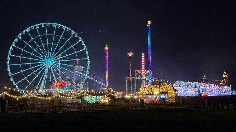 La imagen muestra una vista nocturna de la feria Hyde Park Winter Wonderland en Londres, Reino Unido, con su tradicional rueda de la fortuna gigante iluminada y varias atracciones mecánicas